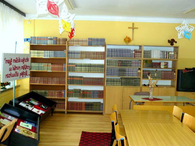 Po lewej stronie znajdują się gabloty wystawowe, po prawej stoliki i krzesła. W tle stoją regały z książkami.