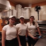 Trzy uśmiechnięte dziewczyny ubrane na biało czarno. Stoją w kuchni. W tle stosy pojemników na pizze.