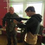 Uczeń trzyma broń w rękach. Żołnierz udziela mu instrukcji