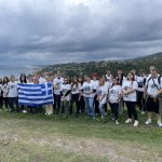 Grupa osób na wybrzeżu trzymają w rękach rozłożoną flagę Grecji.