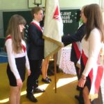Grupa uczniów udekorowanych w szarfy czerwono-białe stoją przy sztandarze. Przekazują sobie sztandar.